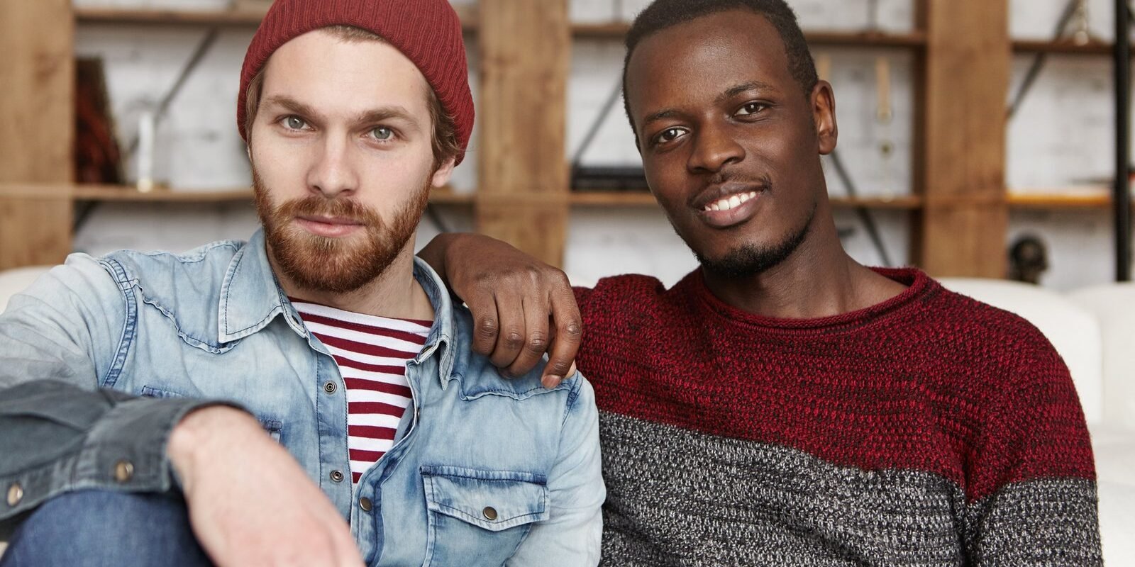 Homosexual love and relationships concept. Interracial gay couple relaxing at cafe: African-American man in sweater holding hand on his stylish bearded Caucasian boyfriend's shoulder in trendy hat Homosexual love and relationships concept. Interracial gay couple relaxing at cafe: African-American man in sweater holding hand on his stylish bearded Caucasian boyfriend's shoulder in trendy hat